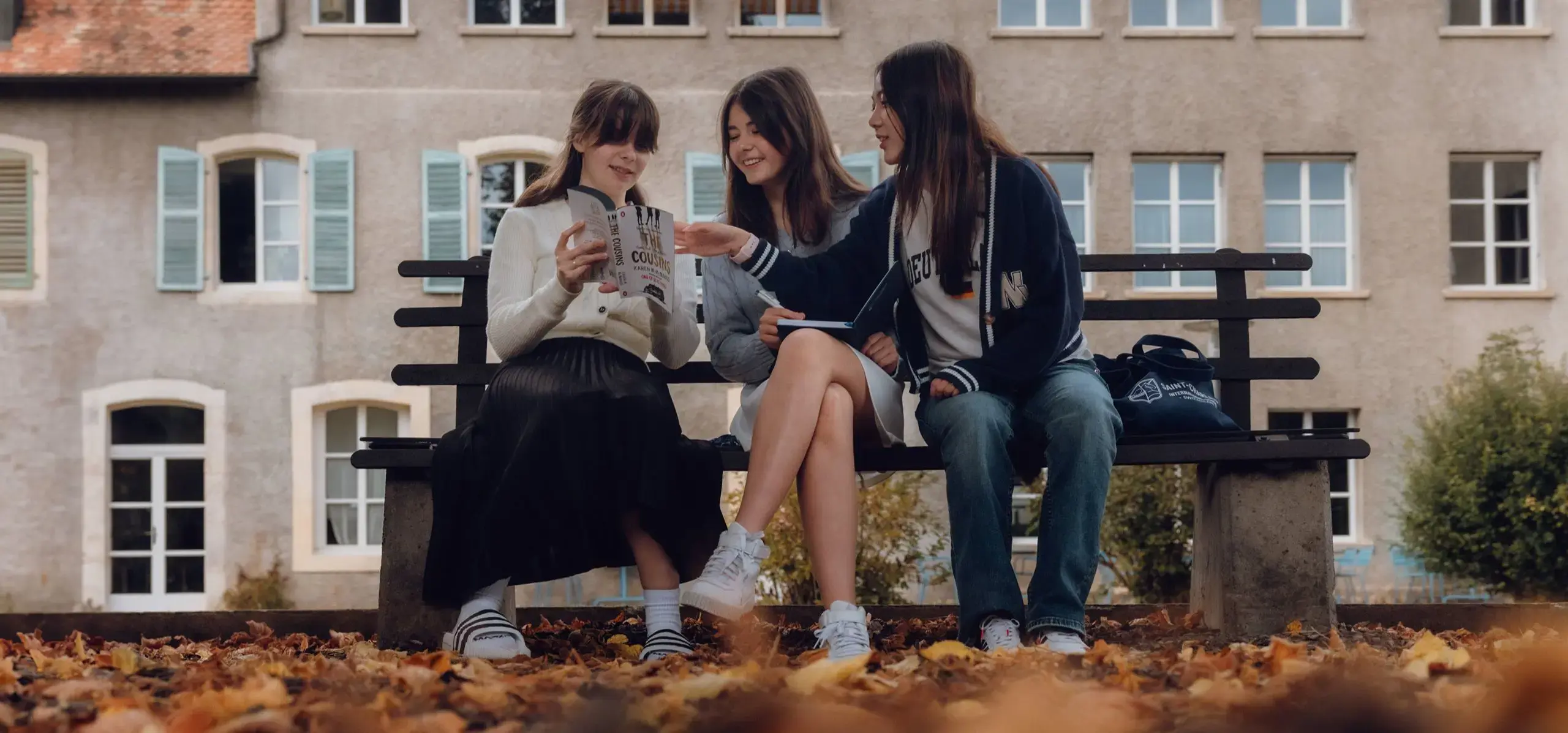 Three students sitting on a bench, discussing the book their friend is reading.